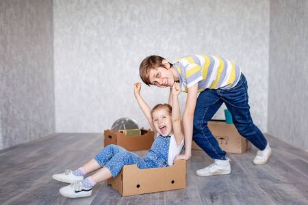 Brother rides sister in a cardboard box. Children play with boxes at home in quarantine. A boy and a girl have moved into a new apartment and are happily playing without toys.の写真素材