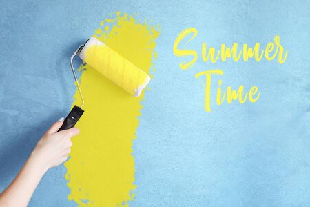 Cropped image of a hand holding a paint roller while painting a house wall in yellow with the inscription summer time. Waiting for summerの写真素材