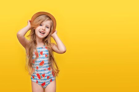 Portrait of a happy child a little blonde girl with long hair in a swimsuit and a hat on a Colored yellow background. The concept of recreation. Summer timeの写真素材