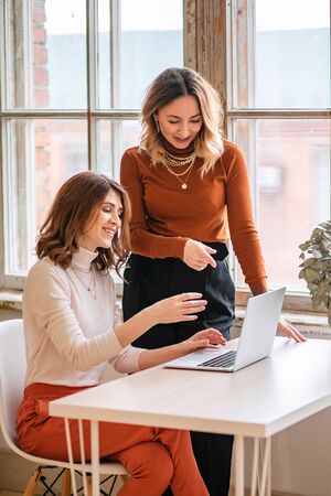 One-on-one meeting.Two young business women sit at a Desk in the office. The girl shows her colleague information on the laptop screen. Women discuss their work online and blog.の写真素材