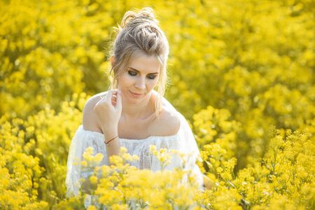beautiful blonde girl with long hair in a rapeseed field in a white dress in a field of yellow flowers blooming in a rapeseed field at sunset or sunriseの写真素材