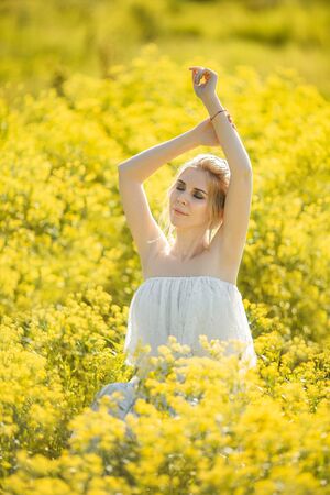 beautiful blonde girl with long hair in a rapeseed field in a white dress in a field of yellow flowers blooming in a rapeseed field at sunset or sunriseの写真素材