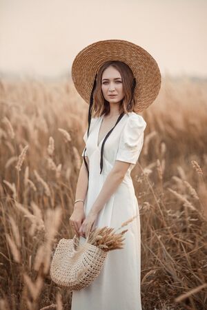 A beautiful Brunette girl in a light white dress and a straw boater hat is holding a straw bag.の写真素材