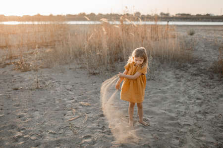 little blonde girl plays on the river in the sand against the sunset skyの写真素材