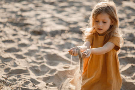 little blonde girl plays on the river in the sand against the sunset skyの写真素材