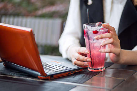 A young female student drinks fresh lemonade while working remotely at a laptop in a cafe in a sunny...