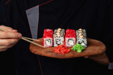 Close-up of the hands of a Man chef in a black uniform holding a wooden tray plate full of sushi wasabi gingerの写真素材