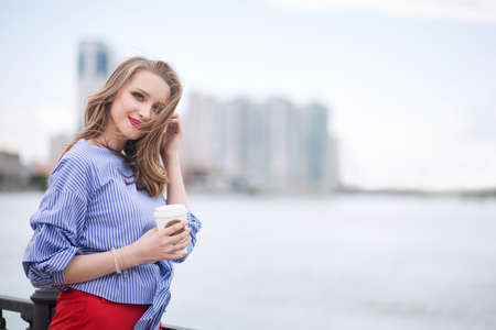 A young beautiful woman is drinking coffee on the city embankment and enjoying a walkの写真素材