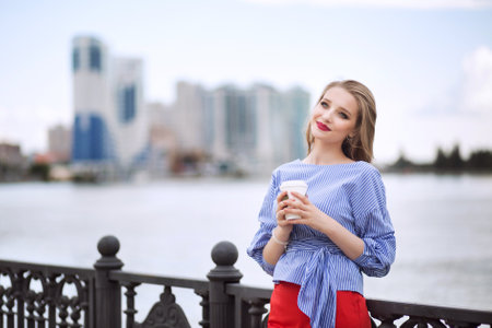 A young beautiful woman is drinking coffee on the city embankment and enjoying a walkの写真素材
