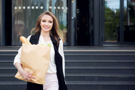 A young beautiful girl with a grocery bag carrying groceries in paper bags comes out of the supermarket outsideの写真素材