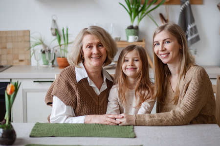 Happy family, elderly grandmother with daughter and granddaughter hugging happily at the table in...