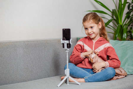 Small blonde blogger girl, an influential person, communicates with subscribers, conducts a live broadcast with a cat, looks at the smartphone screen at home. When taking a selfie, call your friends.の写真素材