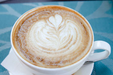 Coffee with foam in a cup is on the table with a brown tablecloth. Coffee house