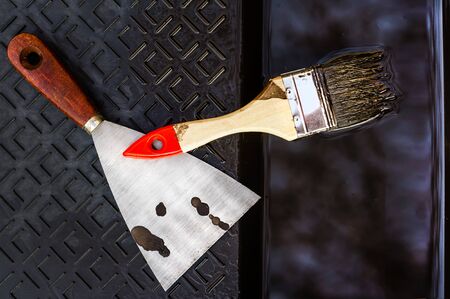Close up of a wooden brush and a shpatel laying in a black roller tray with stains of brown paint