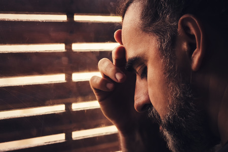 Sad and depressed man holding his head with his hand, standing by the windowの写真素材