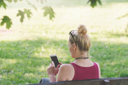 Woman sitting on a park bench in the tree shadow, texting on her phoneの写真素材