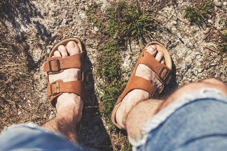 Top view of male legs in brown leather sandals and blue jean shorts, standing on a rocky trailの写真素材