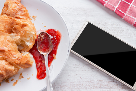 Croissant with jam on white plate and white kitchen table, with red napkin and cell phone beside itの写真素材
