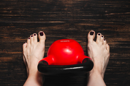 Woman standing bare foot with red kettlebell between her legs on dark wooden floor, view from aboveの写真素材