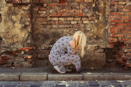 Blond woman in summer dress crouching in front of an old brick wall, hiding her face and cryingの写真素材