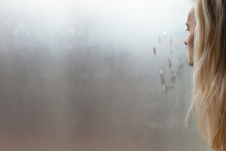 Blonde woman alone, sad and depressed looking through misted window with palm print in front of her faceの写真素材