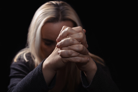 Woman praying with her hands clasped in front of her face, in dark roomの写真素材