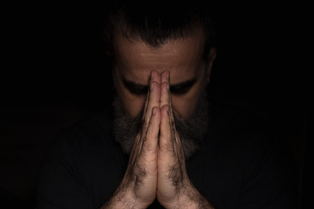 Man bowing his head in prayer with clasped hands in front of his face, in dark room