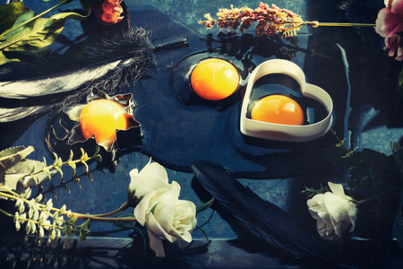 Three raw egg yolks and whites, two in cookie cutters, one spilled on a table, decorated with spring flowers and black feathers for Easter holidayの写真素材