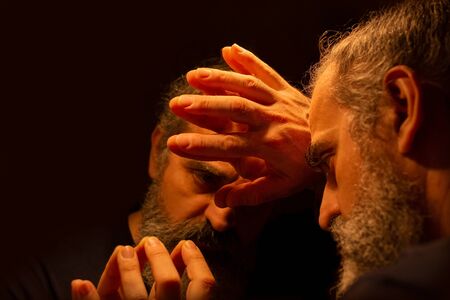 Reflection of bearded man in a dark, holding his head with his hands with painful expression on his...