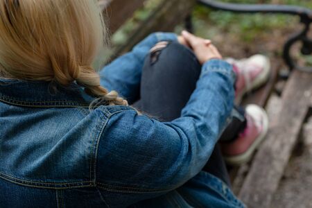 Blonde woman in jeans jacket sitting alone on a park bench, holding her leg with her armsの写真素材