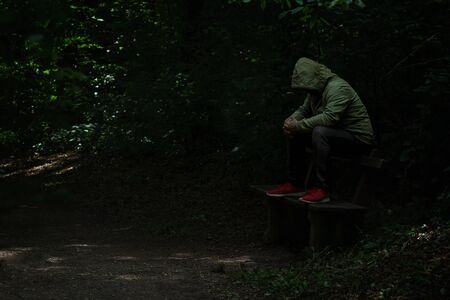 Man sitting on a bench in light spot on forest path, surrounded with darkness, green hoodie covering his face, alone, sad and depressedの写真素材