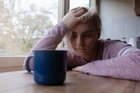 Blonde woman sitting at the kitchen table, looking distraught, holding hand on her head, cup of coffee in front of herの写真素材