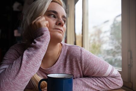 Blonde woman sitting by the window, looking into the distance, cup of coffee on table in front of herの写真素材