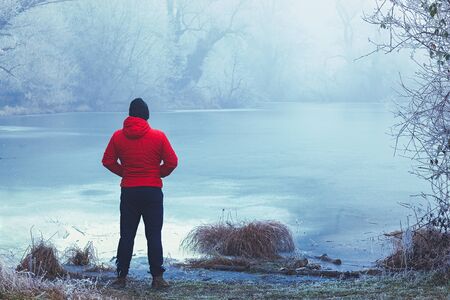 Lonely man in red jacket standing by the frozen lake, looking into the distance, winter landscapeの写真素材