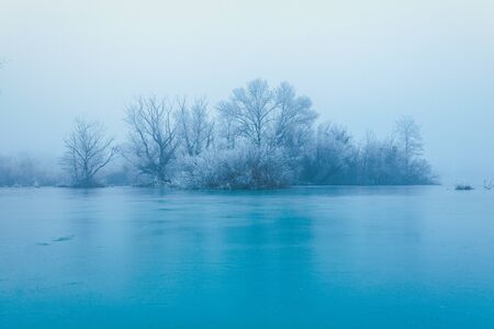 Frozen turquoise lake in misty winter morning, with trees without leaves on a small island across...