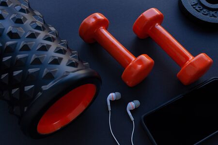 Two orange dumbbells, weight plates, white in ear headphones and bumpy foam roller on gym floorの写真素材