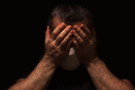 Man with white medical mask on his face, holding his head with his hands, alone and scared in the...