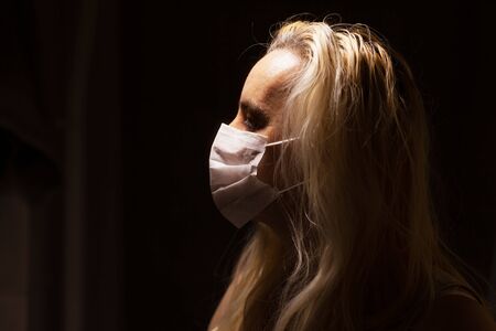 Blonde woman with white medical mask sad and lonely in the dark, with light coming from above,...