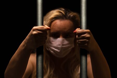 Blonde woman behind the bars wearing white medical mask, holding bars with her hand, alone in dark