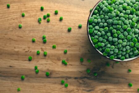 Frozen green peas in a bowl with few scattered on the wooden tableの写真素材