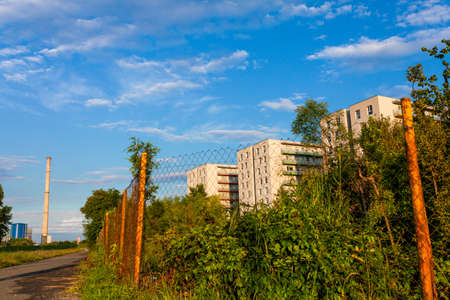 New residential buildings behind lush foliage and wire fence, bright blue sky and heating plant in backgroundの写真素材