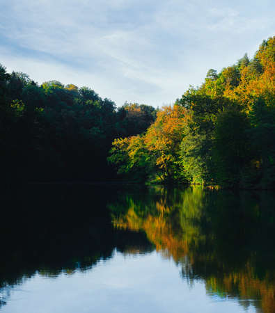 Autumn colors on the trees by the lake, deep shadows and sky reflection in water