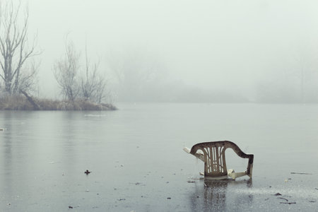 Old, dirty, plastic lawn chair abandoned in frozen lake, foggy and cold dayの写真素材
