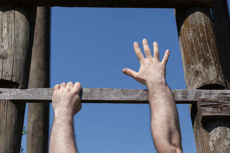 Strong male hands firm gripping  wooden ladder step, one hand reaching up, hanging, blue sky in...