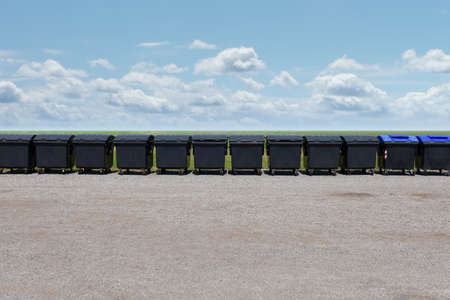 Long row of black garbage containers outside under the blue sky with white clouds, on the gravel