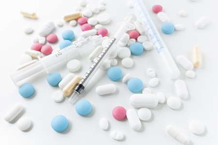 Pile of blue, white, pink and yellow colored pills and capsules with medical syringes with black and...