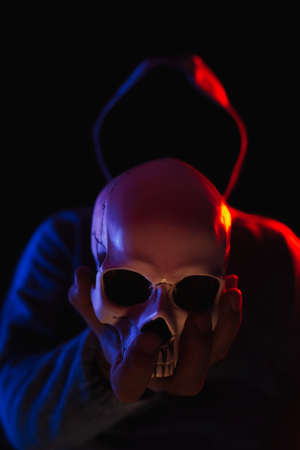 Dark figure in hoodie holding human skull in hand in front, illuminated with red and blue light,...