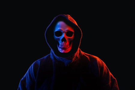 Man with human skull instead of face in dark hoodie illuminated with red and blue lightsの写真素材