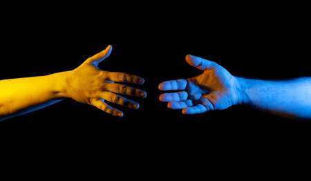 Two hands, male and female, one in yellow one in blue light reaching for each other, black backgroundの写真素材