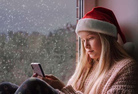 Cute teenage girl in Santa hat using mobile on snowy day during Christmas holidaysの写真素材